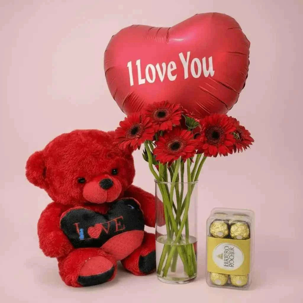زهور جيربيرا حمراء في مزهرية مع شوكولاتة فيريرو روشيه ودمية دب وبالون قلب flowerna